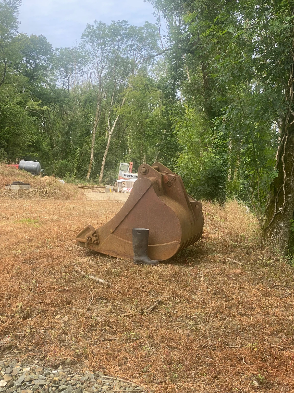 Large detached excavator bucket photographed beside a boot, with machinery behind it.